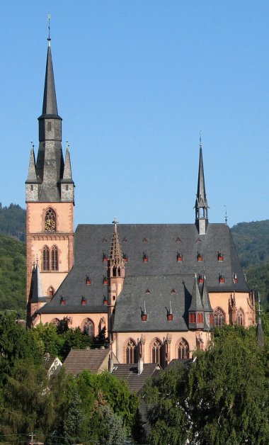 Blick auf die Kirche St. Valentinus und Dionysius Kiedrich und der Michaelskapelle mit dunklen Dachziegeln, rötlichen Steinen und dunkelroten Fensterumrandungen und Ziersteinen vor Bäumen und einem blauem Himmel ohne Wolken