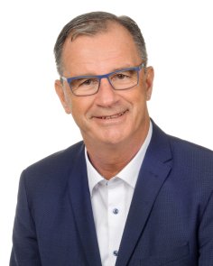 Portrait von Bürgermeister Winfried Steinmacher mit blauer Brille, braunen Augen, dunklen Haaren, blauem Sakko mit weißem Hemd mit hellblauen Knöpfen, lächelnd vor einem weißen Hintergrund.  