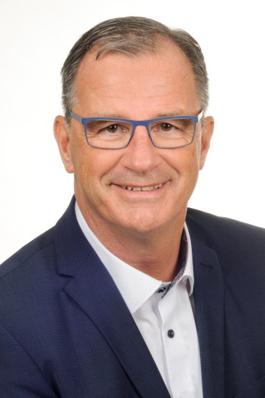 Portrait von Bürgermeister Winfried Steinmacher mit blauer Brille, braunen Augen, dunklen Haaren, blauem Sakko mit weißem Hemd mit hellblauen Knöpfen, lächelnd vor einem weißen Hintergrund.  