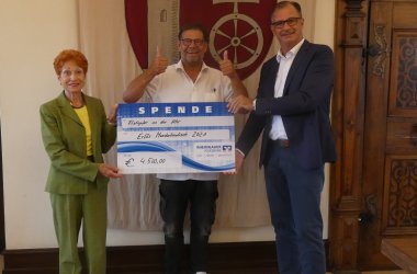 Frau Monika Alber steht neben Bäckermeister Stefan Dries, der die Daumen nach oben hält. Daneben steht Bürgermeister Steinmacher, der mit Frau Albert den großen blau-weißen Spendenscheck der Rheingauer Volksbank über 4.500,00 EUR in den Händen hält.  