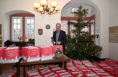 Bürgermeister Winfried Steinmacher steht im Anzug mit einer Weinflasche in der Hand neben einem Tisch mit rot, weißen Adventstüten, neben einem einem geschmückten Weihnachtsbaum und vor weiteren Adventstüten im Rathaussaal. 