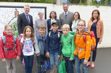 Sechs Schulkinder der John-Sutton-Schule im Vordergrund, hinten dran Staatssekretär Dr. Manuel Lösel, Bürgermeister Winfried Steinmacher, Brigitte Pietsch, Schulleiterin Petra Kelm-Kühne und zwei weitere Frauen vor einer weißen Mauer mit dem Mosaik der John-Sutton-Schule, erkennbar die Burg Scharfenstein mit roter Fahne. 
