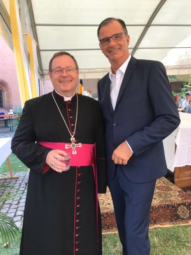 Zusammentreffen Winfried Steinmacher im blauen Anzug mit weißem Hemd mit Brille und Bischof Georg Bätzing mit Brille. Seine Kasel ist schwarz mit pinkem breitem Gürtel und pinken Knöpfen. Um den Hals trägt er eine lange Kette mit einem Kreuz.