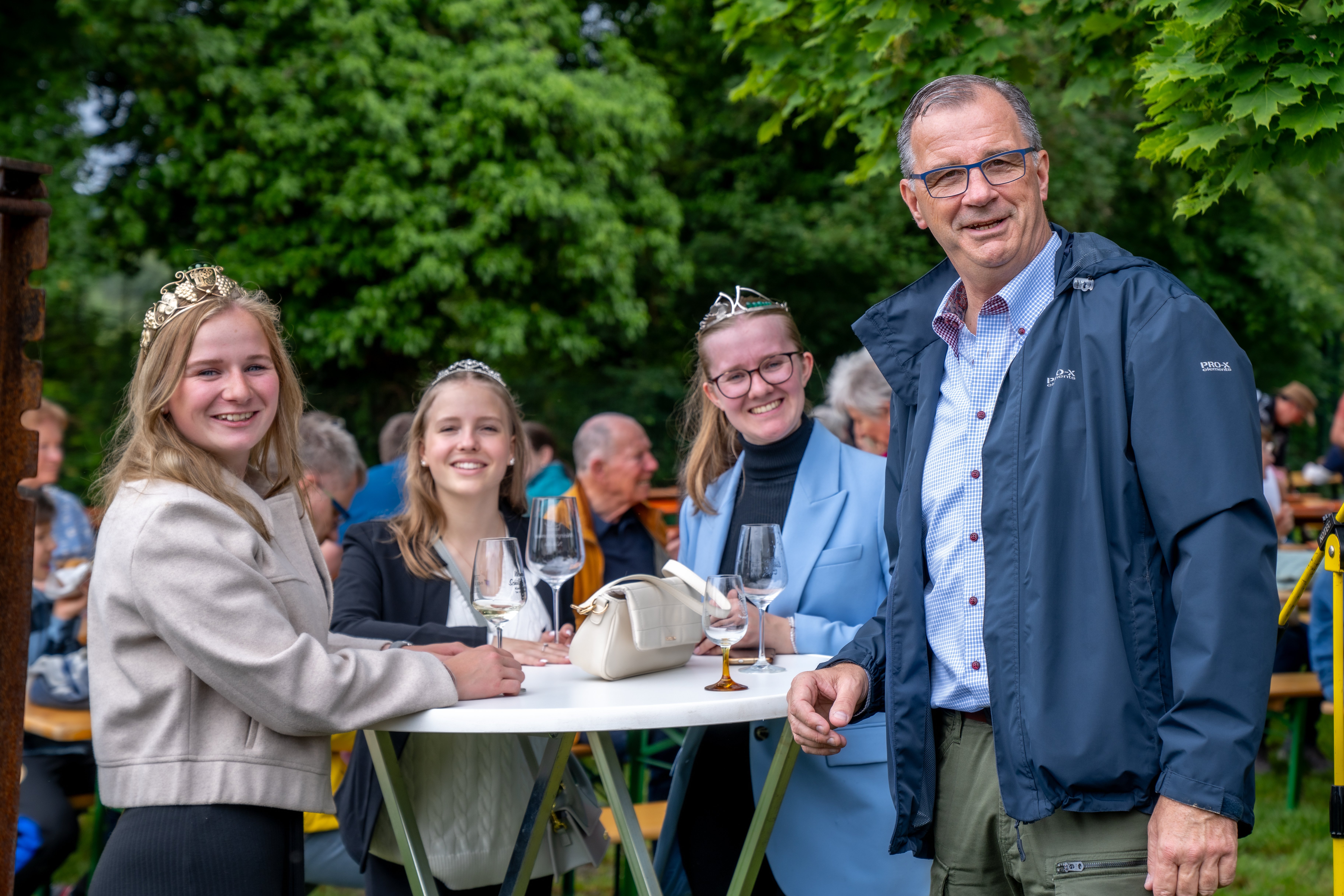 Die Weinkönigin und die Weinprinzessinen sowie Bürgermeister Steinmacher stehen an einem Bistrotisch mit Weingläsern vor belaubten Bäumen.