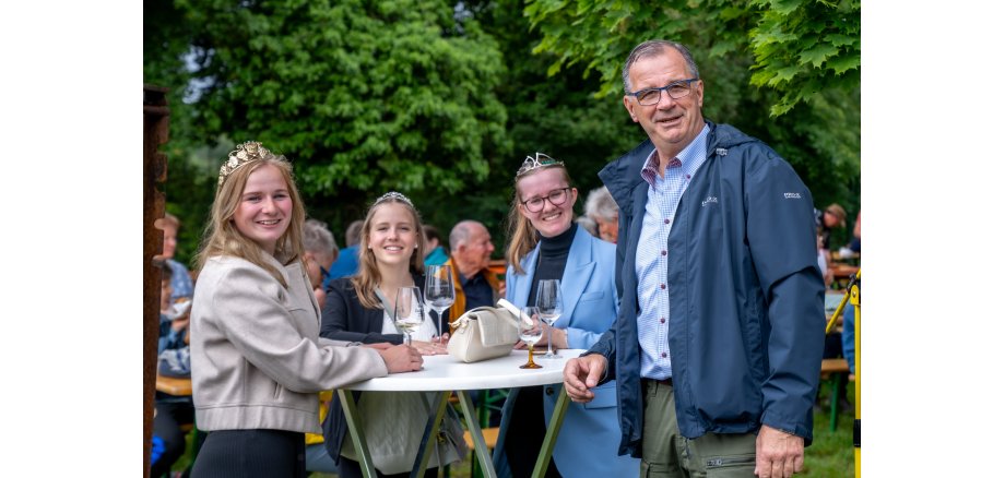 Die Weinkönigin und die Weinprinzessinen sowie Bürgermeister Steinmacher stehen an einem Bistrotisch mit Weingläsern vor belaubten Bäumen.