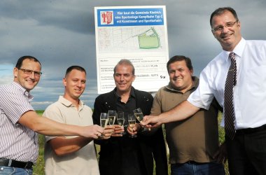 Vier Männder Heiko Preußer, Alexander von Alkier, Raimond Gülin, Swen Domröse und Bürgermeister Winfried Steinmacher, die mit Sektgläsern anstoßen und vor einem Bau-Schild stehen. Im Hintergrund der Himmel mit grauen Wolken. 