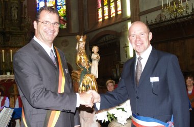 Bürgermeister Winfried Steinmacher im dunklen Anzug mit Krawatte mit einer Schärpe in schwarz, rot, gold gibt Monsieur Lopez in dunkelblauem Anzug und Krawatte mit Schärpe in  blau, weiß, roter, die Hand. Im Hintergrund die Statue von der Jungfrau Maria mit dem Jesukind und  dem Chorgestühl, Kronleuchtern, Kerzen und bunten Kirchenfenstern. 