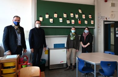 In einem Klassenraum der Kiedricher Grundschule stehen vor einer Tafel mit 16 Bildern, von links Bgm. W. Steinmacher, Johannes Scholl, Schulleiterin Petra Kelm-Kühne sowie eine Klassenlehrerin, alle mit Corona-Gesichts-Maske und ein weißes, rechteckiges  Entkeimungsgerät.  