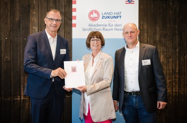 Bürgermeister Steinmacher, Priska Hinz und Christian Paff stehen im Anzug mit der Annerkennungsurkunde vor einem Banner von Hessen mit dem Text Land hat Zukunft. 