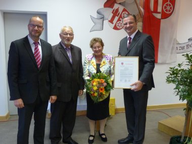 Harald Rubel und Hubertus Harras neben Wilma Scholl und Bürgermeister Winfried Steinmacher. Die Herren im dunklen Anzug und Frau Scholl im schwarzen Rock und buntem Blazer mit einem gelb, orangenem Blumenstrauß in der Hand. Herr Bürgermeister Steinmacher hält die Urkunde im goldenem Bilderrahmen. AN der Wand hängt die Kiedricher Fahne in rot und weiß und auf der Wand befindet sich das gemalte Kiedricher Wappen in rot und weiß. 