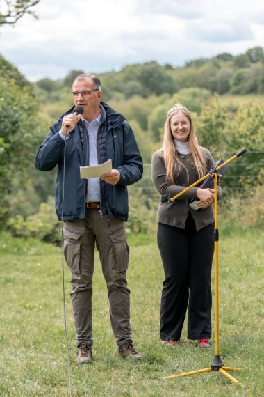 Auf einer Wiese steht ein Mann mit blauer Jacke, hellblauem Hemd und dunkelgrüner Hose. Er hat einen Zettel in der Hand und hält ein Mikrofon vor seinen Mund. Daneben steht eine Frau mit Brille und langen blonden Haaren und einer goldenene Krone auf dem Kopf. Sie trägt eine dunkelgrüne Jacke und eine schwarze Hose. Im Vordergrund ist ein gelber Mikrofonständer. Der Hintergrund ist verschwommen mit einem bewölktem Himmel und Baumkronen.