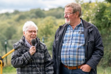 Eine Frau mit schwarz-grau karrierter Jacke, einer roten Brille und weißen, kurzen Haaren hat ein Mikrofon in der Hand. Neben ihr steht ein Mann mit einem braunem Gürtel, einem blau-weiß-gelb karriertem Hemd und einer dunklen Jacke. Er trägt ebenso eine Brille, hat einen weißen Bart und hellbraune Haare.