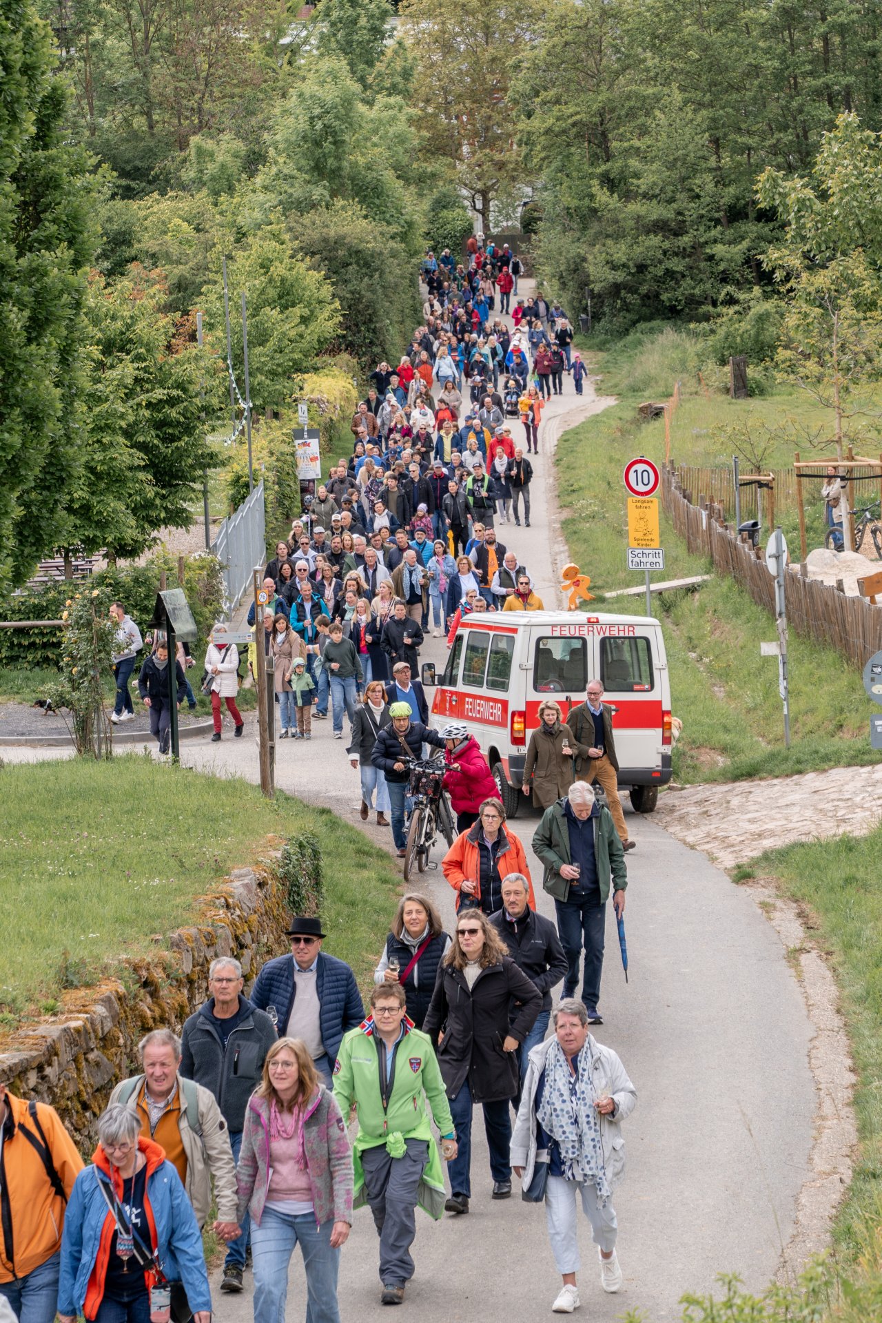 Auf einem asphaltierten Weg laufen viele Personen mit Jacken und einem Weinglas in der Hand. Vorn ist Wiese zu erkennen und im hinteren Bereich viele Bäume. Ein Kleinbus der Feuerwehr in weiß-rot ist ebenso auf dem Weg.  