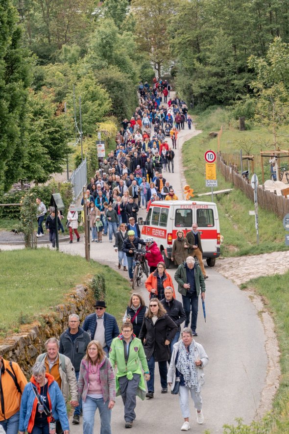 Auf einem asphaltierten Weg laufen viele Personen mit Jacken und einem Weinglas in der Hand. Vorn ist Wiese zu erkennen und im hinteren Bereich viele Bäume. Ein Kleinbus der Feuerwehr in weiß-rot ist ebenso auf dem Weg.  