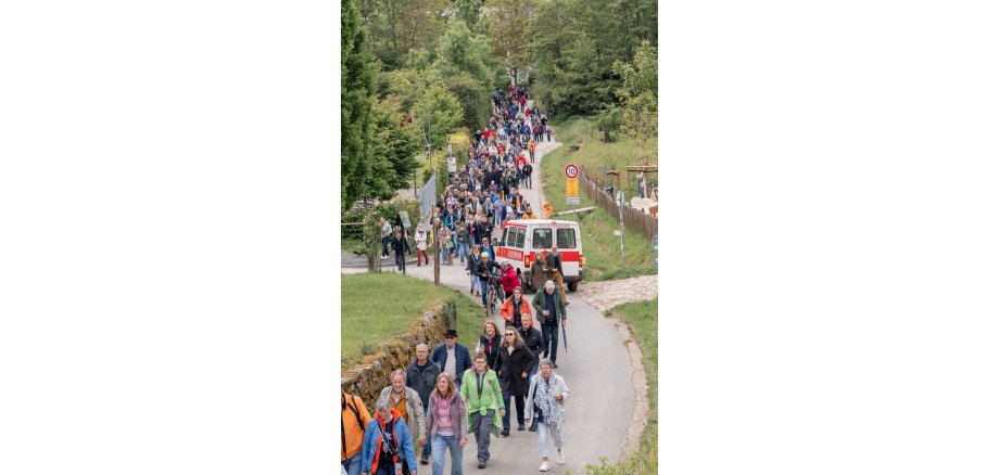 Auf einem asphaltierten Weg laufen viele Personen mit Jacken und einem Weinglas in der Hand. Vorn ist Wiese zu erkennen und im hinteren Bereich viele Bäume. Ein Kleinbus der Feuerwehr in weiß-rot ist ebenso auf dem Weg.  