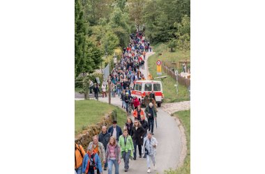 Ehepaar-Treffen 2026 Auf einem asphaltierten Weg laufen viele Personen mit Jacken und einem Weinglas in der Hand. Vorn ist Wiese zu erkennen und im hinteren Bereich viele Bäume. Ein Kleinbus der Feuerwehr in weiß-rot ist ebenso auf dem Weg.