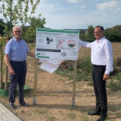 Bürgermeister Steinmacher und weiterer Mann vor dem Hinweisschild, was auf den Grünschnittplatz entsorgt werden darf auf Erdfläche vor einem Baum.  