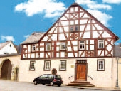 Weißes Fachwerkhaus mit braunen Balken und weißen Sprossenfenstern und Steintreppe mit einem dunklem davor parkendem Auto.