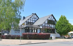 Blick auf ein weißes Fachwerkhaus mit braunen Balken und grünen Bäumen, Hecken und blauem Himmel