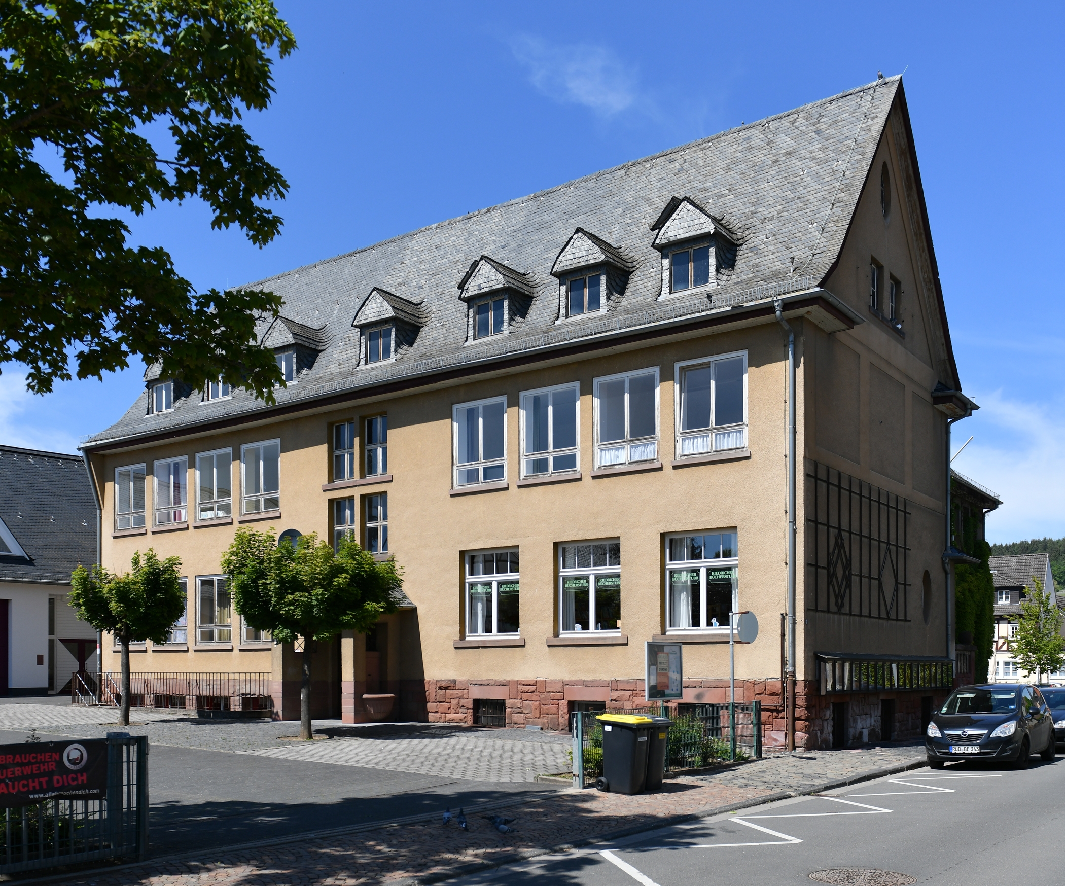 Alte Schule Kiedrich Beiges Gebäude mit Fenstern und Dachfenstern in dem mit schwarzen Ziegeln bedeckten Dach, In dem Gebäude befand sich früher die Schule von Kiedrich. Ebenso sieht man den Hof mit Bäumen und ein Teil der Straße mit einem parkendem Auto.
