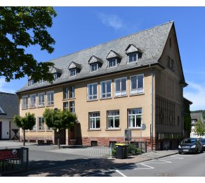 Beiges Gebäude mit Fenstern und Dachfenstern in dem mit schwarzen Ziegeln bedeckten Dach, In dem Gebäude befand sich früher die Schule von Kiedrich. Ebenso sieht man den Hof mit Bäumen und ein Teil der Straße mit einem parkendem Auto. 