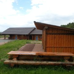 Blick auf das Holzhaus mit blauen Türen und einem großen an einer Seite offeneer Holzuntersteller mit Kiesboden. Auf der Wiese steht eine aus Baumstämmen gefertigte Bank mit Tisch. Links von dem Blockhaus ist ein Basketballkorb aus Metall und ein aspahltierter Platz zu erkennen. Im Hintergrund sind belaubte Bäume. 