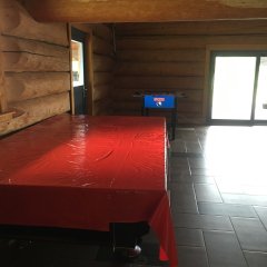Innenansicht, Wand aus Baumstämmen, Boden mit grauen, hellgrauen und schwarzen, rechteckigen Fliesen. Links ist eine Tür und geradeaus die zweiflüglige Terrassentür. In dem Raum steht ein blauer Tisch-Kicker und im Vordergrund ein mit einer roten Plastiktischdecke zugedeckter Billardtisch. 