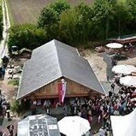 Luftbild mit Blick auf das Blockhaus mit der Kiedricher Fahne in rot und weiß und weiß aufgeklappte Sonnenschirme und Pavillon und vielen Leuten. Im Hintergrund belaubte Bäume und ein Feldweg und ein Feld. 