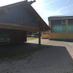 Blick auf das Holzblockhaus mit einer Terrassentür und Schieferplatten am Giebel. Man sieht Wiese und eine asphaltierte Terrasse und rechts ist ein gelbes Gebäude mit Glasstreifen.