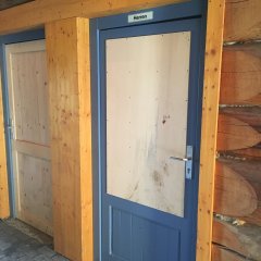 Gepflasterter Boden und Blick auf die Wand aus Baumstämmen und den beiden WC-Türen mit blauem Rahmen und Türen aus Holz mit dem Beschriftungs-Schild "Herren".