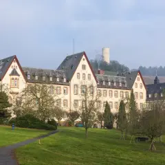 HInter einer Wiese mit mehreren Laubbäumen und einem Weg, steht ein mehrgeschössiges beiges Gebäude mit drei Dachgibeln. Rechts davon ist ein weiteres, mehrgeschössiges beiges Gebäude und im Hintergrund ist die Burgruine Scharfenstein auf einem Berg erkennbar. Der Himmel ist weiß.