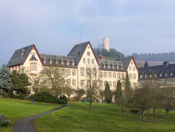 HInter einer Wiese mit mehreren Laubbäumen und einem Weg, steht ein mehrgeschössiges beiges Gebäude mit drei Dachgibeln. Rechts davon ist ein weiteres, mehrgeschössiges beiges Gebäude und im Hintergrund ist die Burgruine Scharfenstein auf einem Berg erkennbar. Der Himmel ist weiß.