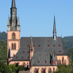 Blick auf die Kirche St. Valentinus und Dionysius Kiedrich und der Michaelskapelle mit dunklen Dachziegeln, rötlichen Steinen und dunkelroten Fensterumrandungen und Ziersteinen vor Bäumen und einem blauem Himmel ohne Wolken