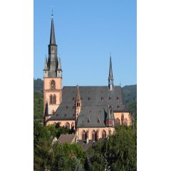 Blick auf die Kirche St. Valentinus und Dionysius Kiedrich und der Michaelskapelle mit dunklen Dachziegeln, rötlichen Steinen und dunkelroten Fensterumrandungen und Ziersteinen vor Bäumen und einem blauem Himmel ohne Wolken