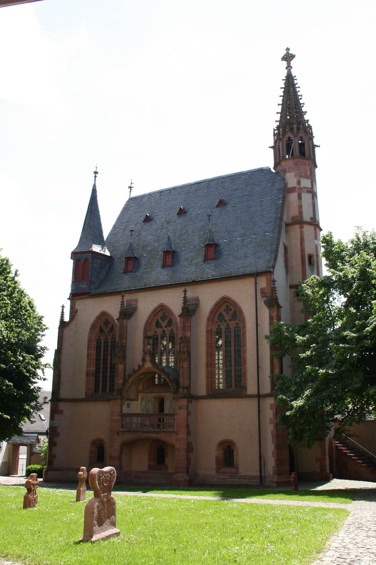 Blick auf die Michaelskapelle mit dunklen Dachziegeln, rötlichen Steinen und dunkelroten Fensterumrandungen und Ziersteinen drei Türmen und einem Erker sowie einer Steintreppe zwischen Bäumen und hinter einer Wiese mit drei sandsteinfarbenen Grabsteinen und einem Weg aus Pflastersteinen vor einem weissen Himmel.