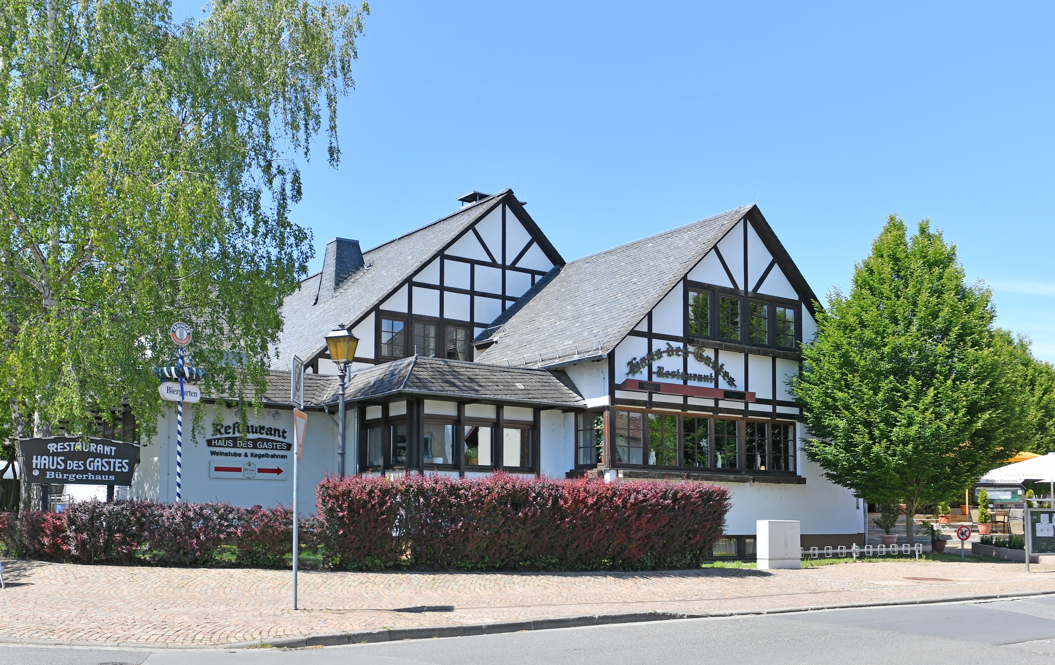 Blick auf ein weißes Fachwerkhaus mit braunen Balken und grünen Bäumen, Hecken und blauem Himmel