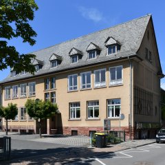 Beiges Gebäude mit Fenstern und Dachfenstern in dem mit schwarzen Ziegeln bedeckten Dach, In dem Gebäude befand sich früher die Schule von Kiedrich. Ebenso sieht man den Hof mit Bäumen und ein Teil der Straße mit einem parkendem Auto. 