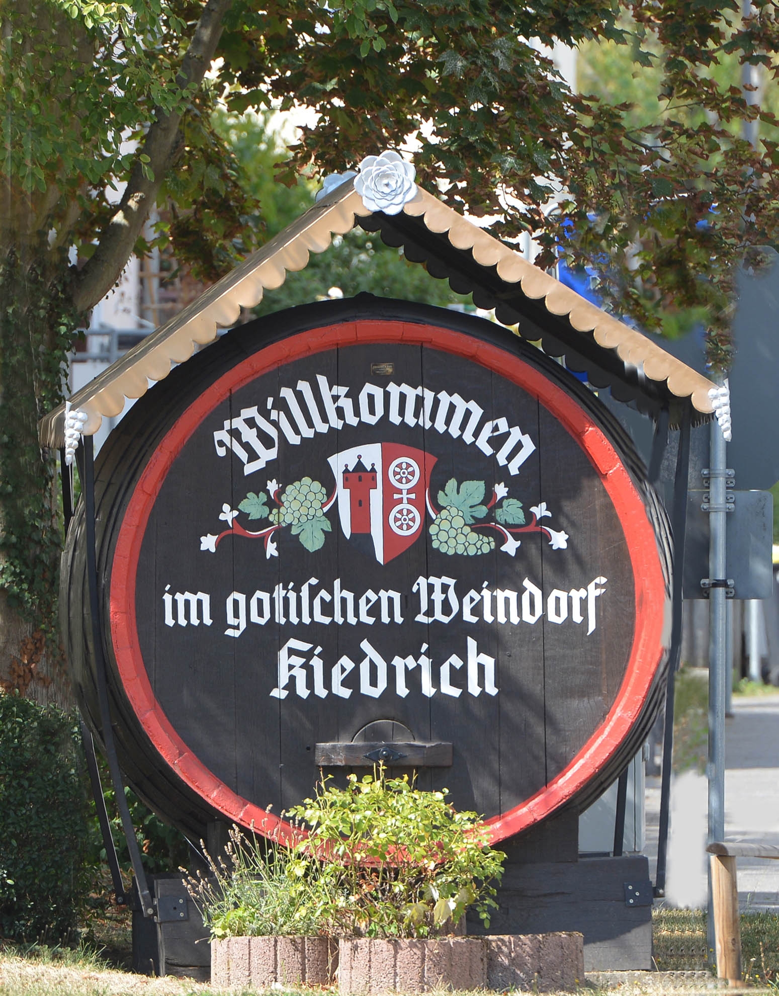 Altes Weinfass aus Holz mit Begrüßungstext und Kiedricher Wappen mit Trauben und roter Umrandung sowie einem Schutzdach.