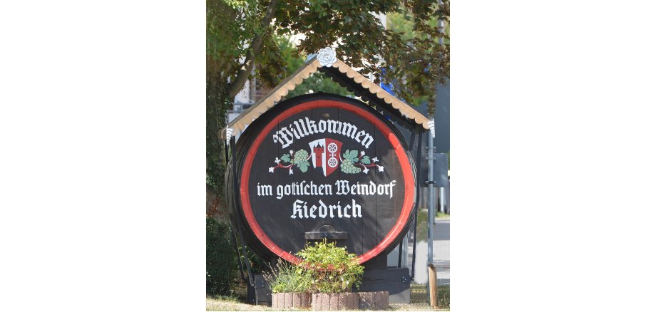 Begrüßungsfass Kiedrich Altes Weinfass aus Holz mit Begrüßungstext und Kiedricher Wappen mit Trauben und roter Umrandung sowie einem Schutzdach.