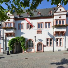 Kiedricher Rathaus, weisses Gebäude mit Sandsteinfensterrahmen, zwei Erkern, dem Eingang und einer Fahne mit Kiedricher Wappen in rot und weiss sowie einem mit grünen Blättern umrankten Torbogen.