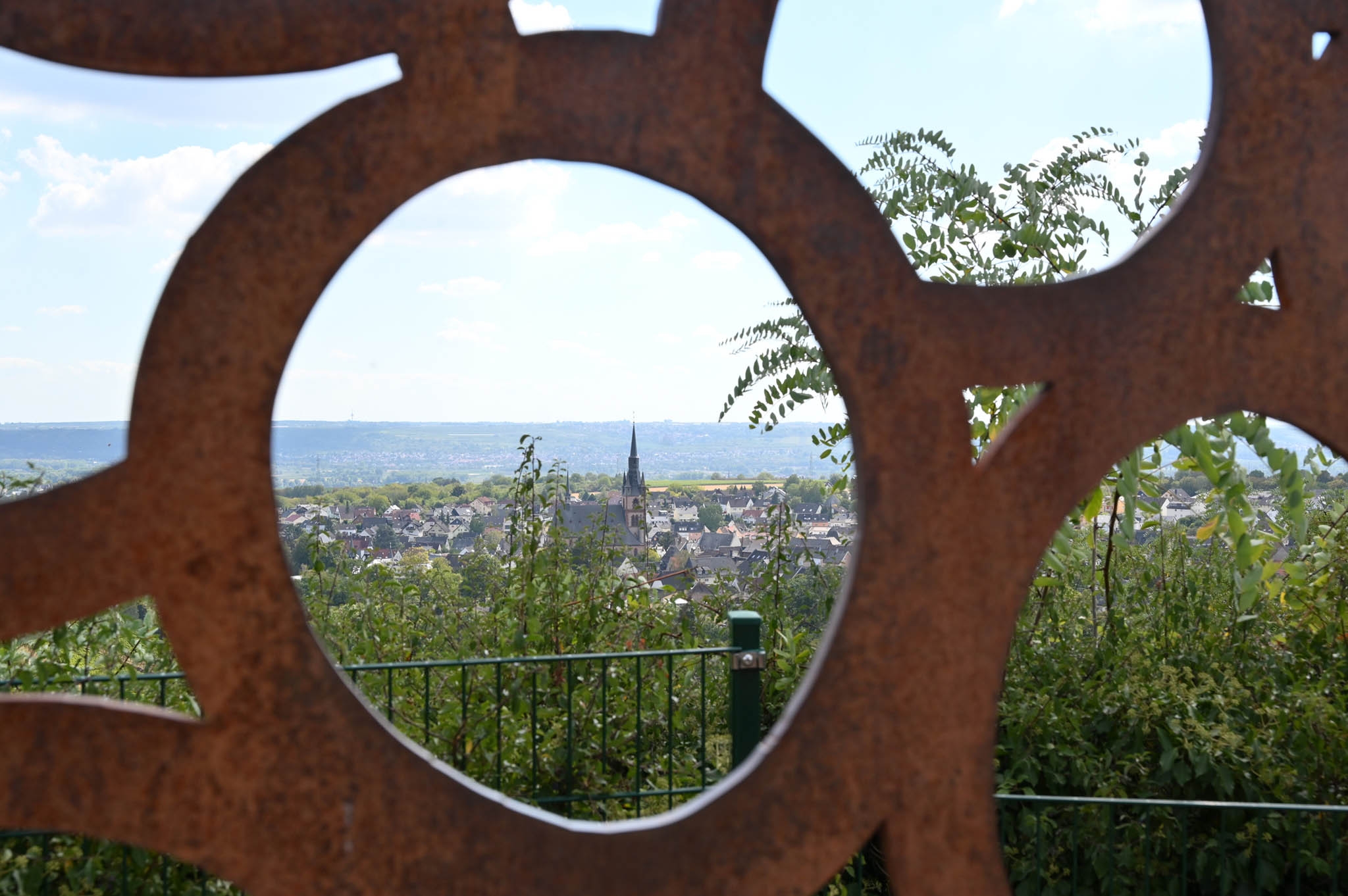 Blick auf Kiedrich durch die Skulptur mit Weintrauben mit Häusern, Bäumen und der Kirche St. Valentinus und Dionysius 
