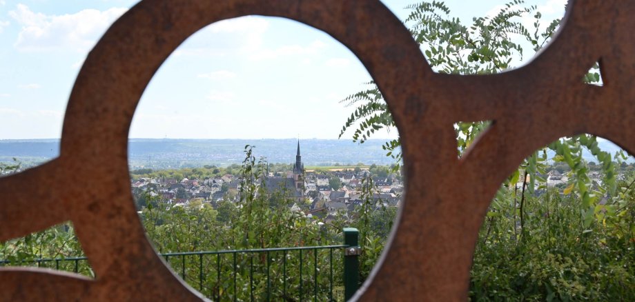 Blick auf Kiedrich durch die Skulptur mit Weintrauben mit Häusern, Bäumen und der Kirche St. Valentinus und Dionysius 