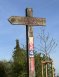 An einem viereckigen Holzpfosten ist ein rechteckiges Schild in blau mit blauem Pfeil und einer weißen 4 und darüber ein rechteckiges Schild in rot mit rotem Pfeil und einer weißen 3 und darüber ein rechteckiges Schild in gelb mit gelbem Pfeil und einer weißen 2. Oben ist aus Holz ein Pfeil, der nach links zeigt und mit zwei braunen Kriesen, die sich überschneiden und in roten Buchstaben steht "Zum Weinberg der Ehe".