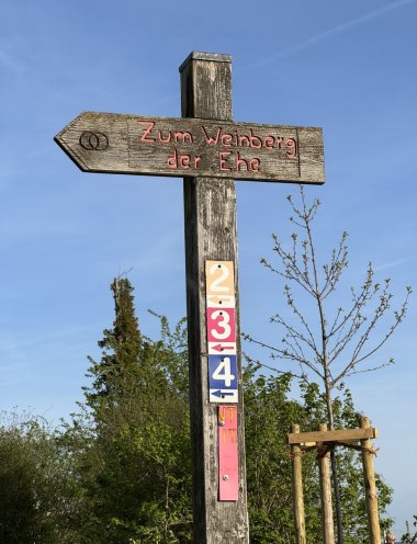 An einem viereckigen Holzpfosten ist ein rechteckiges Schild in blau mit blauem Pfeil und einer weißen 4 und darüber ein rechteckiges Schild in rot mit rotem Pfeil und einer weißen 3 und darüber ein rechteckiges Schild in gelb mit gelbem Pfeil und einer weißen 2. Oben ist aus Holz ein Pfeil, der nach links zeigt und mit zwei braunen Kriesen, die sich überschneiden und in roten Buchstaben steht "Zum Weinberg der Ehe".