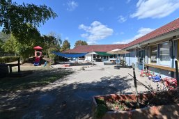 Kita Hickelhäusje Blick auf das Gebäude des Kindergartens Hickelhäusje. Im Vordergrund der Hof mit blauem Sonnensegel, Sandkasten, einer Rutsche, mehrere Dreiräder einem großen Sandkasten, ein bepflanzter Blumenkasten und Bäume.