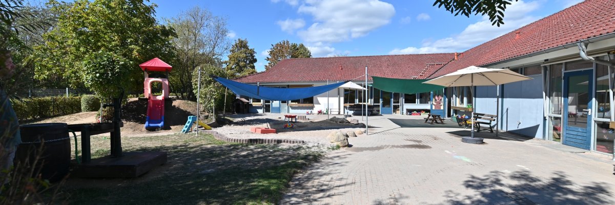 Blick auf das L-förmige Gebäude des Kindergartens Hickelhäusje. Im Vordergrund der Hof mit blauem und grünem Sonnensegel, ein beiger aufgeklappter Sonnenschirm, ein großer Sandkasten, zwei Rutschen und einer Matschbahn aus Holz mit Brunnenpumpe und Holzfass.