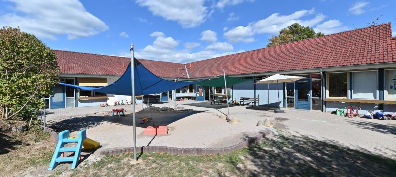 Blick auf das L-förmige Gebäude des Kindergartens Hickelhäusje. Im Vordergrund der Hof mit blauem und grünem Sonnensegel, einem beigen aufgespannten Sonnenschirm,  mehrere Dreiräder, einem großen Sandkasten, eine kleine Rutsche und Rasen sowie Bäume. 