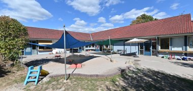 Blick auf das L-förmige Gebäude des Kindergartens Hickelhäusje. Im Vordergrund der Hof mit blauem und grünem Sonnensegel, einem beigen aufgespannten Sonnenschirm,  mehrere Dreiräder, einem großen Sandkasten, eine kleine Rutsche und Rasen sowie Bäume. 