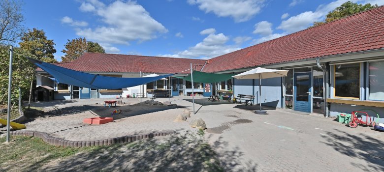 Blick auf das L-förmige Gebäude des Kindergartens Hickelhäusje. Im Vordergrund der Hof mit blauem und grünem Sonnensegel, einem beigen aufgespannten Sonnenschirm,  mehrere Dreiräder und einem großen Sandkasten. 