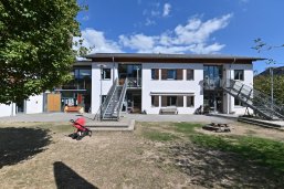 Katholische Kindertagesstätte St. Valentin Weißes Gebäude mit zwei Metalltreppen und Sand und Wiese zum Spielen im Vordergrund.